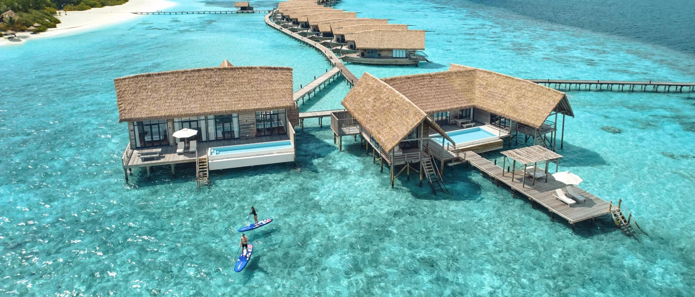 Two people paddle board in a clear turquoise lagoon, with thatched overwater villas with wooden decks, jetties and private pools and a white-sand beach to the left
