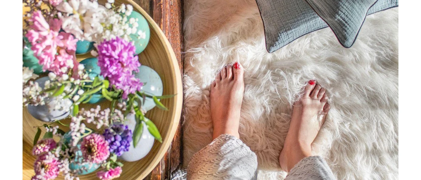 Woman with red painted toenails sinks her feet into a white fur rug next to a table with flowers