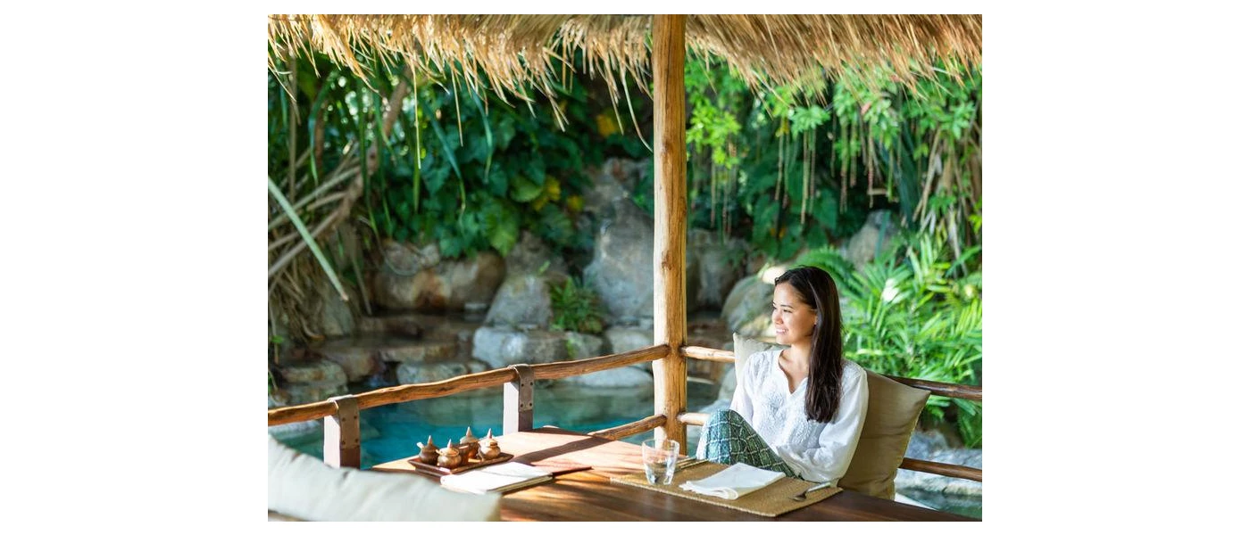 Woman in a white top smiles out at the tropical greenery from a restaurant table in an open-sided cabana