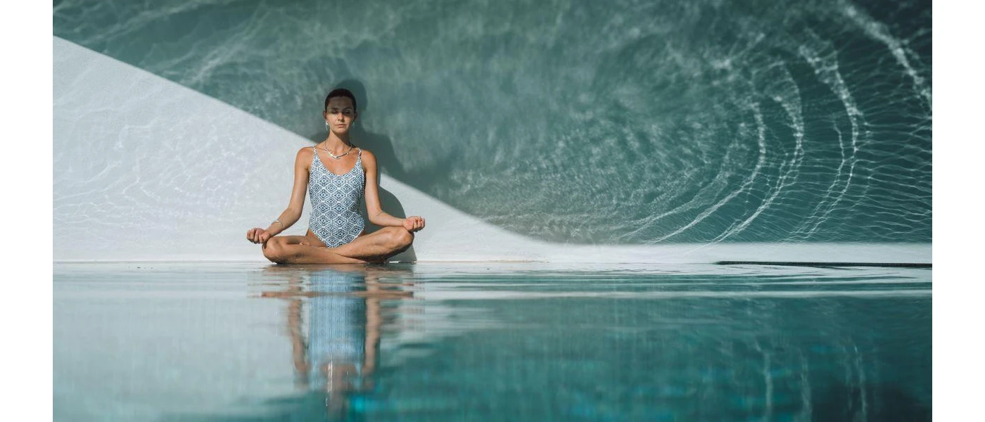 Woman in blue patterned swimsuit sits cross legged with her eyes closed next to a pool with a wave mural on the wall next to her
