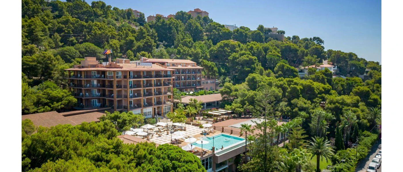 Exterior of Palasiet Wellness Clinic & Thalasso, a multi-storey hotel surrounding by tropical greenery, with an outdoor terrace and swimming pool under a sunny blue sky