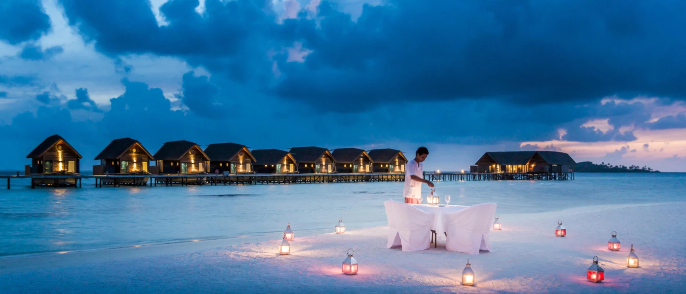 Staff member in white lights a lantern on a table set on the beach, with lanterns all around and overwater villas in the background