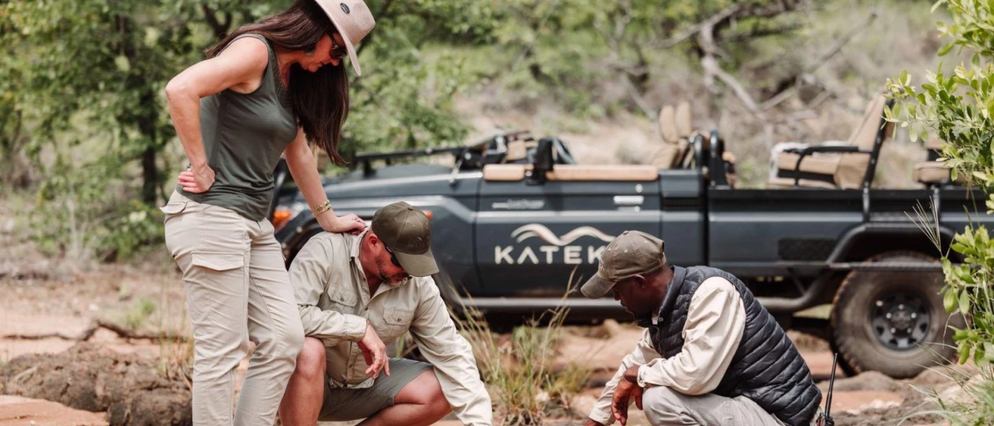 Male and female in camo safari attire look down at the ground as a game drive professional shows them something