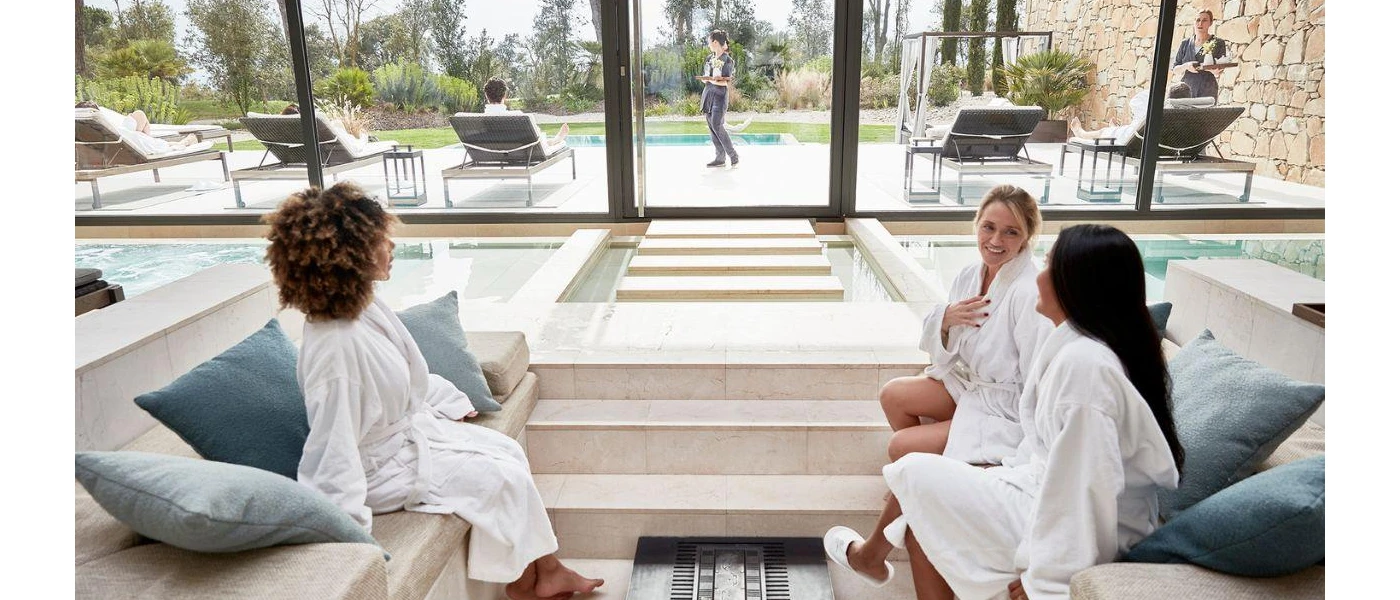 Three woman in white robes sit by an indoor pool with floor-to-ceiling windows overlooking an outdoor pool and gardens
