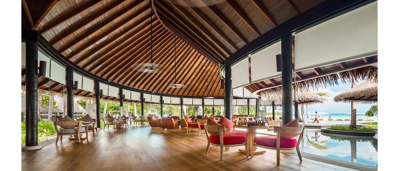 Wooden pavilion with open sides and ceiling fans, furnished with red chairs and wooden tables, one with a chess board