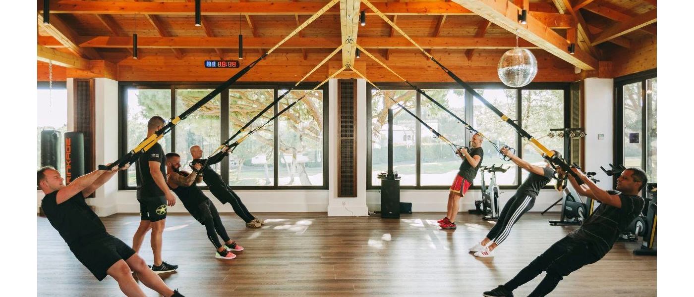 Group in active wear take part in a TRX class with ropes hanging from a wood-beamed ceiling in a studio with floor-to-ceiling windows