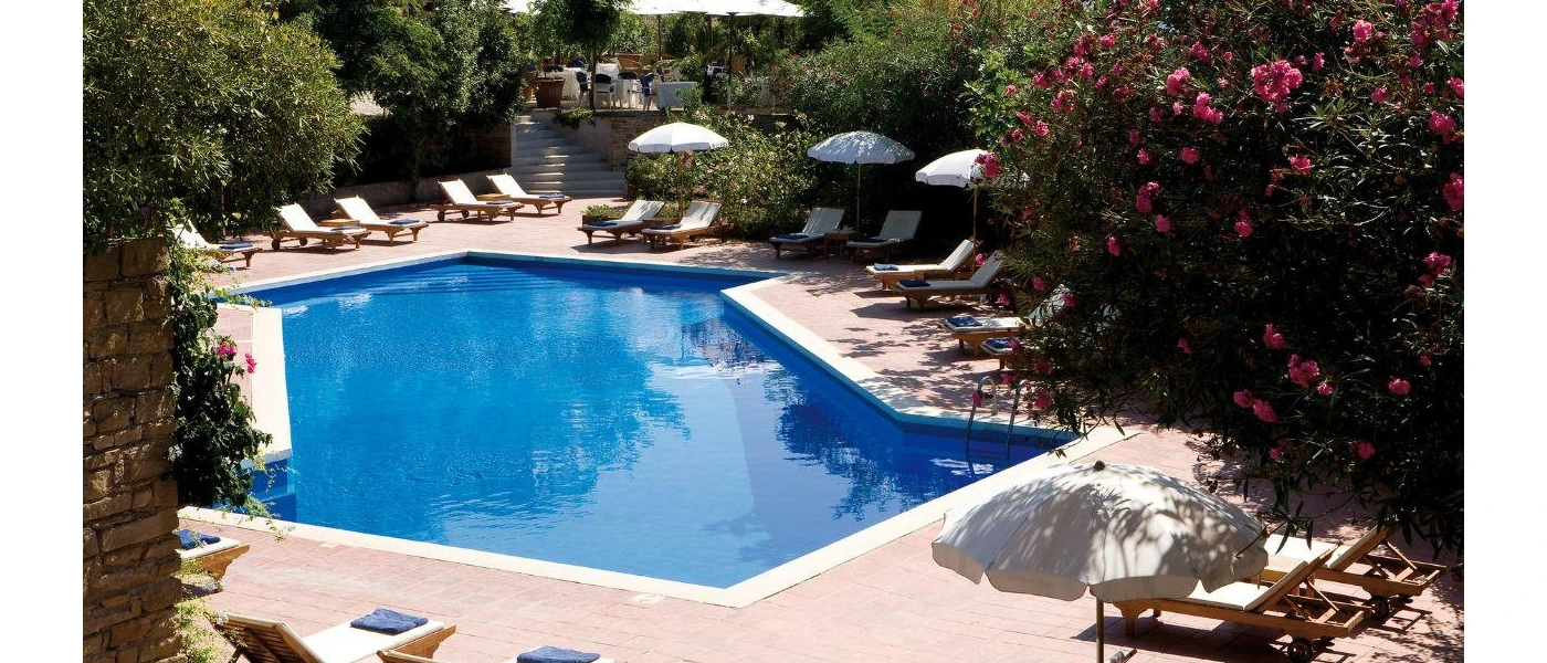 Angular swimming pool surrounded by wooden, white-cushioned loungers and parasols and a thick cocoon of greenery in a sunny terrace