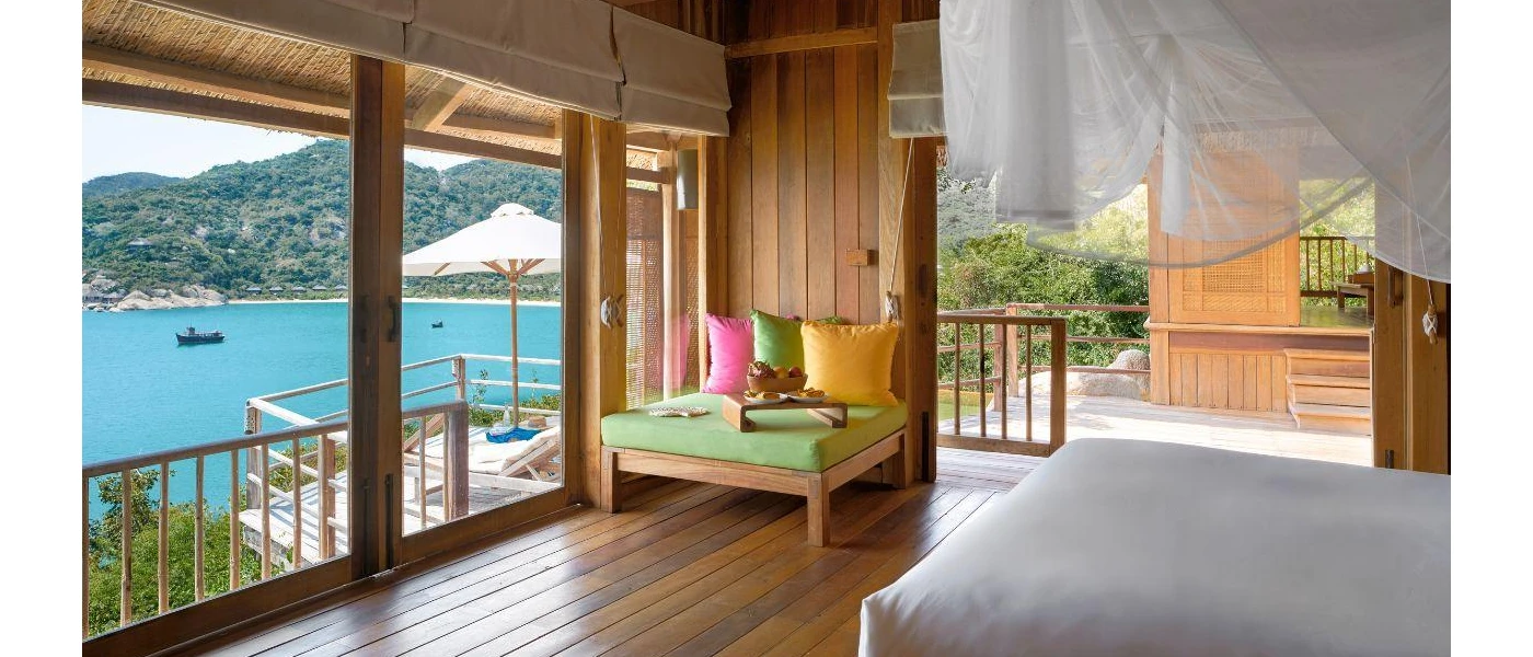Ocean-facing bedroom with white bedding and drapes, wooden flooring and walls, and a wooden chair with green, pink and yellow cushions 