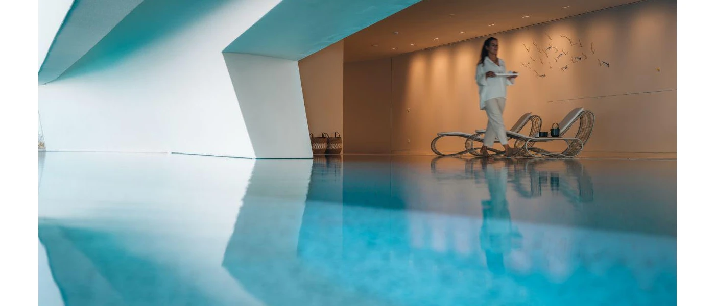 Woman in white walks past an indoor pool past a wall with an art sculpture and softly lit by spotlights