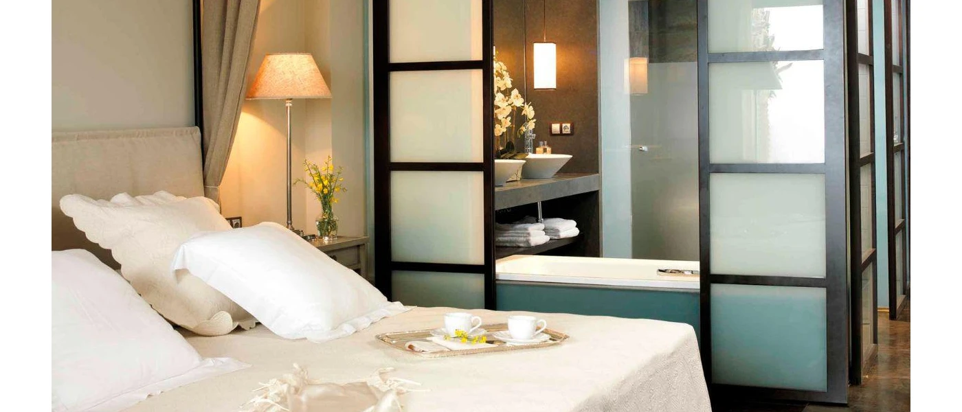 Hotel room with crisp white bedding, sliding black partition doors opening to a bathtub and two sinks