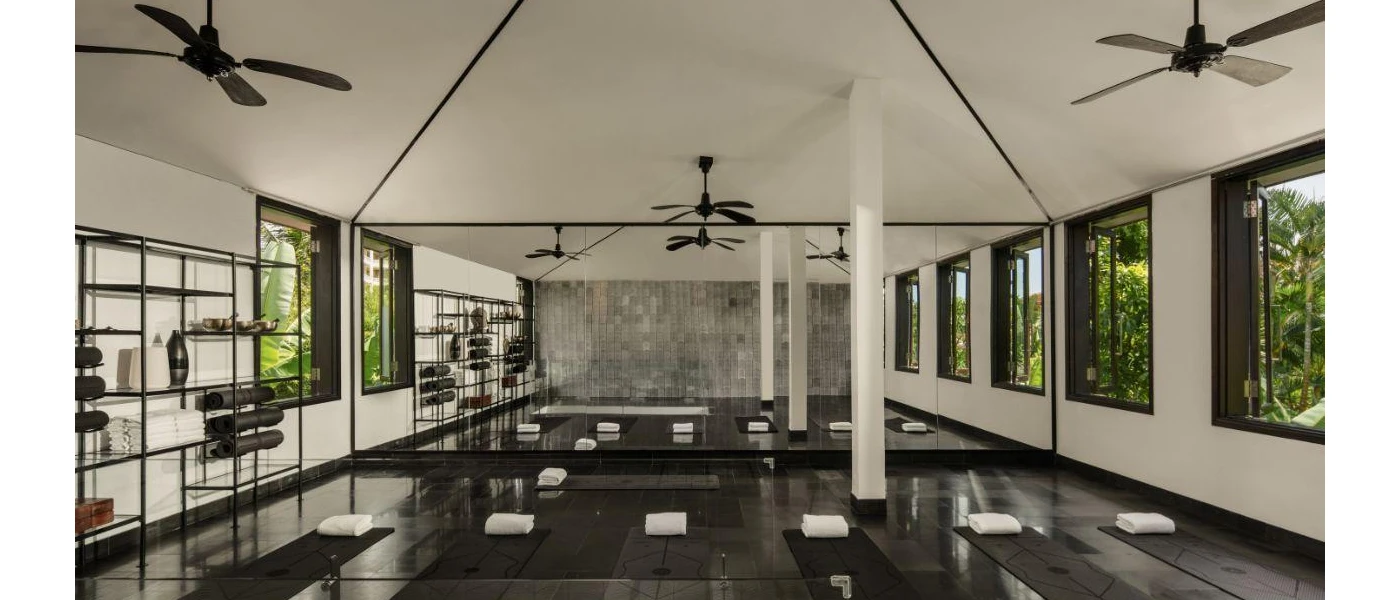 Airy studio with ceiling fans, white walls and dark-wood floors, set up for a class with black yoga mats and white towels