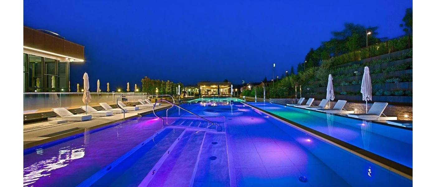 Swimming pool surrounded by white umbrellas and loungers, lit up under a darkening sky
