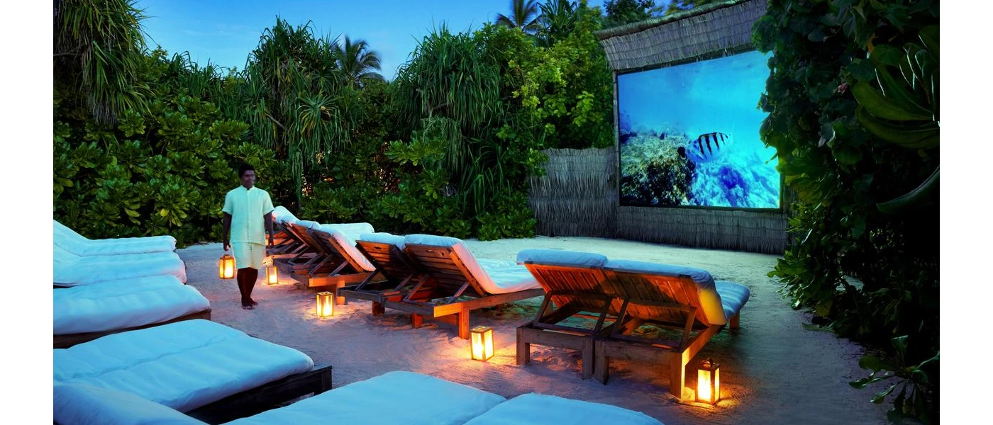 Male staff members carries lanterns to lay with others among white-cushioned daybeds in front of a large screen in an open-air cinema set-up
