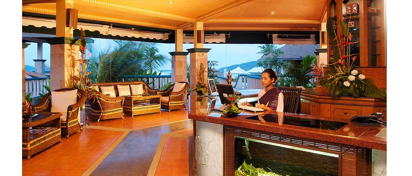 Receptionist in authentic Thai dress stands behind a desk in a traditionally designed reception area, with tiled floors, rattan chairs, dark-wood panelling and a view to the hillsides outside
