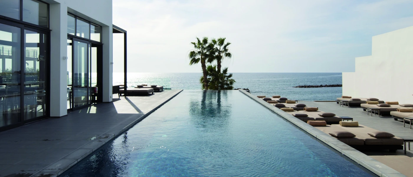 Infinity pool looking over palm trees and the ocean next to a contemporary glass-windowed white building and a line of cushioned daybeds