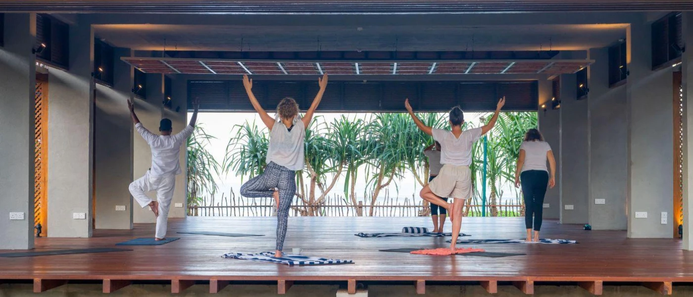 Group on blue and white towels practice yoga in a wooden, open-sided pavilion