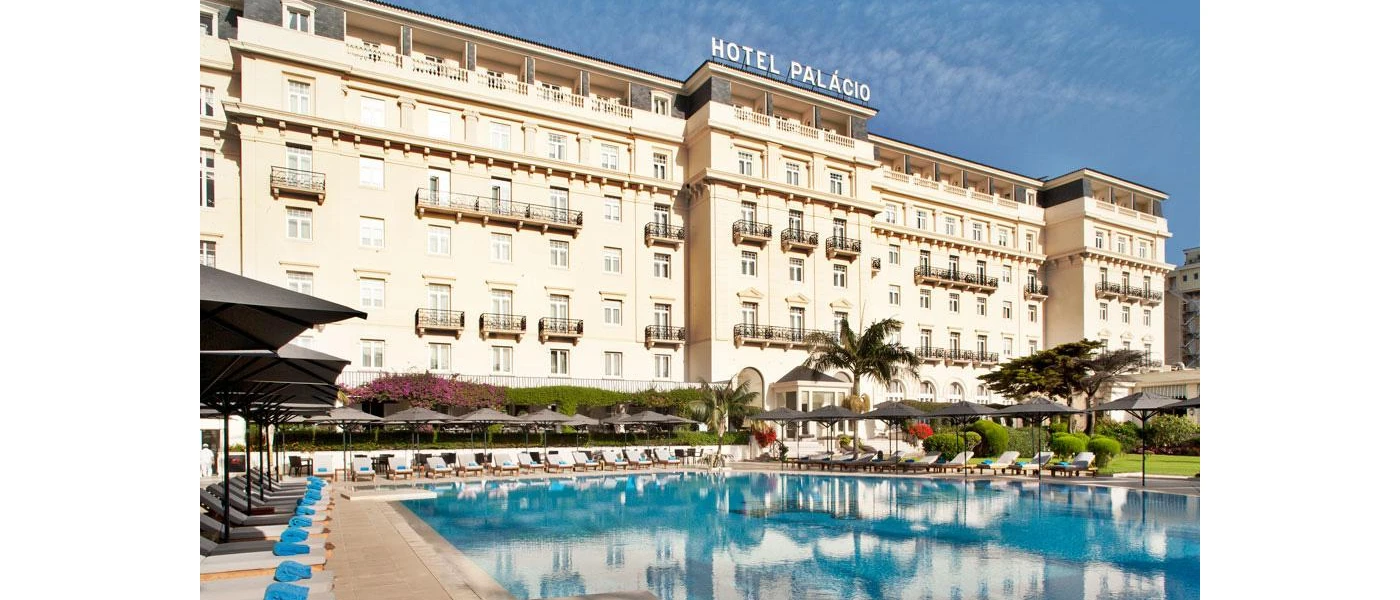 Palacio Estoril's grand, 1930s facade with white walls, black wrought-iron balconies and a swimming pool under a blue sky