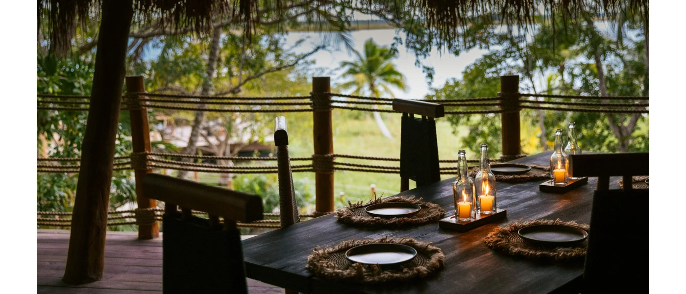 Outdoor restaurant terrace with a lagoon view, thatched roof, wooden tables and rattan placemats with candle centrepieces
