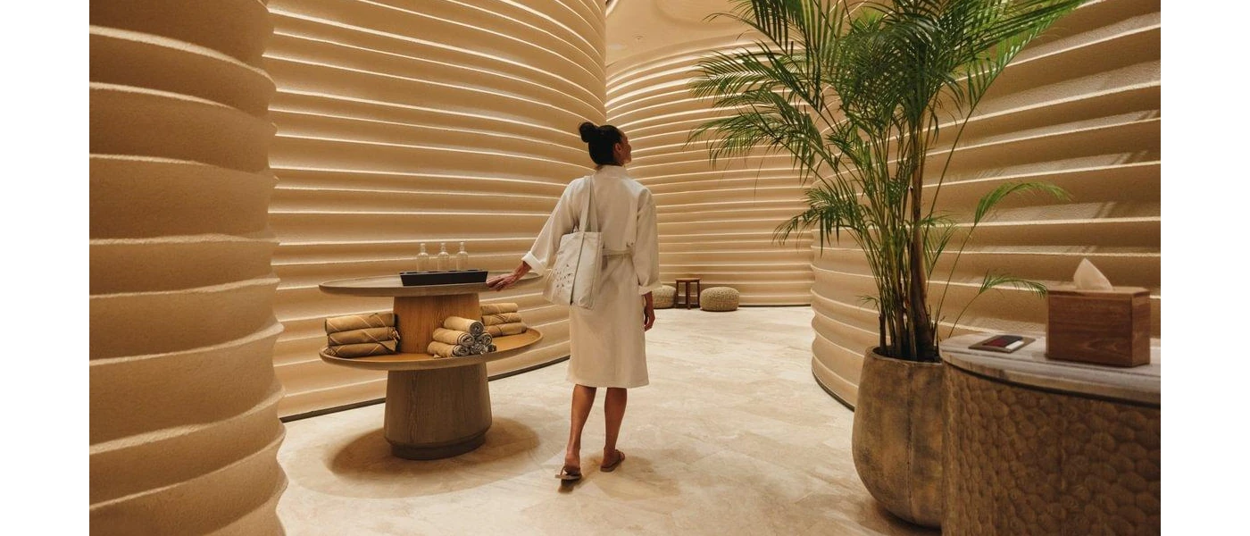 White-robed woman walking into a spa corridor with curved walls, a potted plant and a table stacked with towels and clear bottles