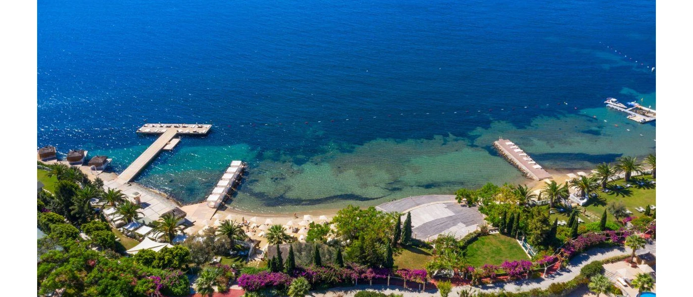 Bougainvillea-framed pathway fronted by lawns and trees overlook a sand and shingle beach and turquoise waters 