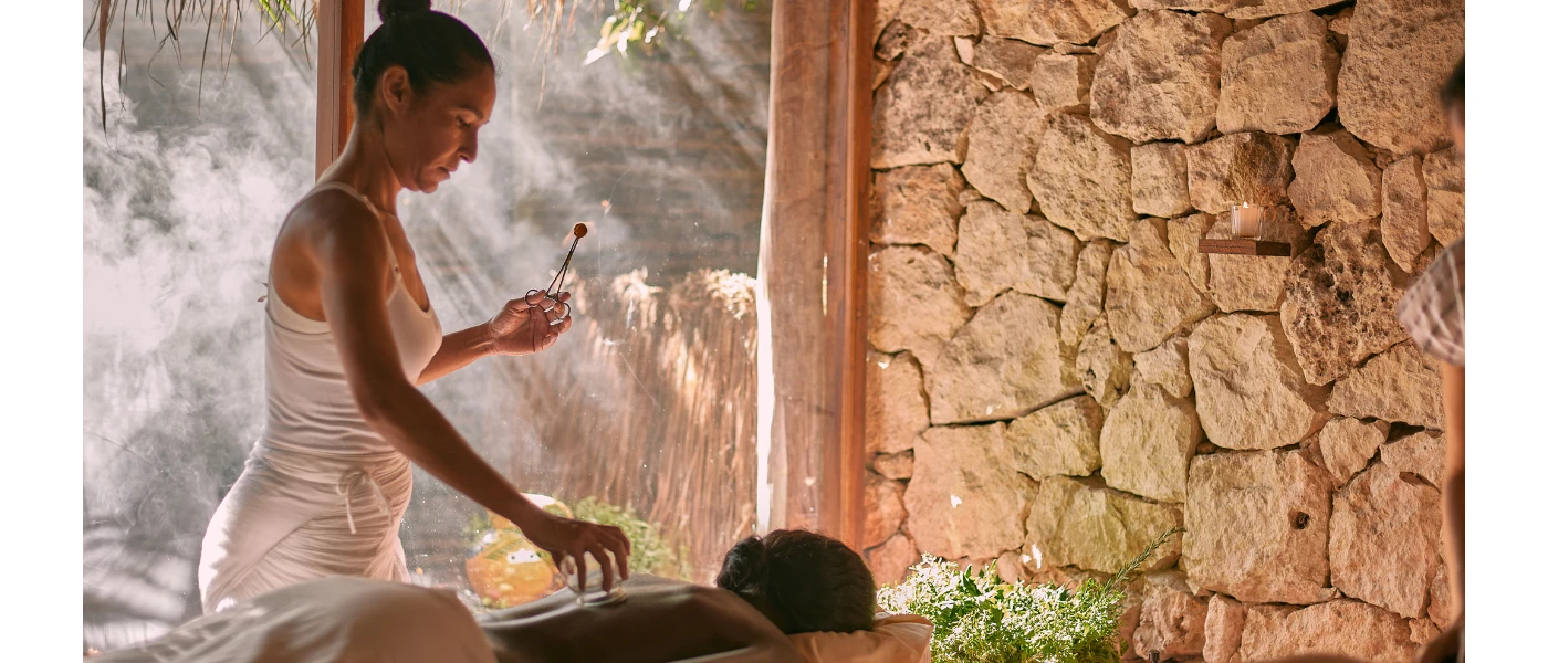 Therapist in white carries out a spa treatment as a woman lies on her front in a room with a cobbled stone wall