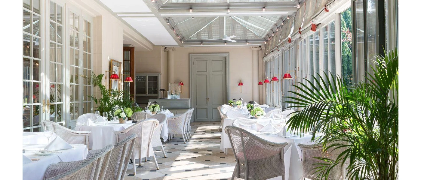 Tables with crisp white tablecloths lined up in sunny conservatory area, with green plants, a patterned floor and red hanging lights