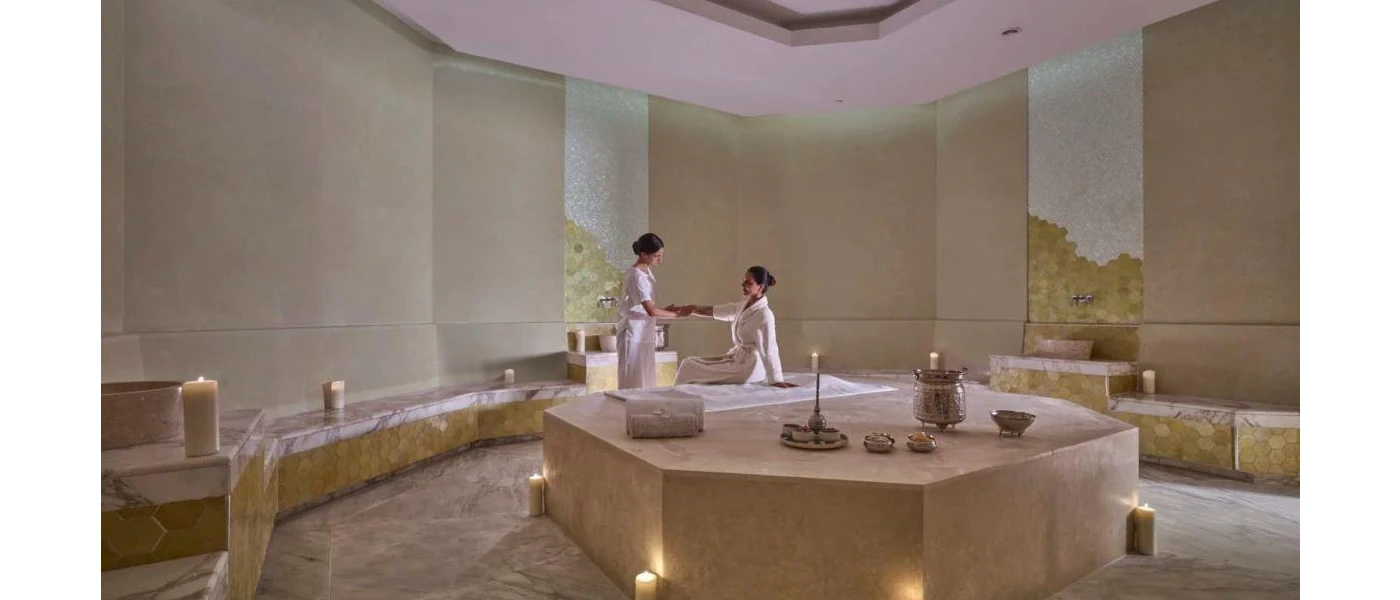 White-robed woman has her arm massaged by a therapist in a candlelit marble-cloaked room with cream walls and floors