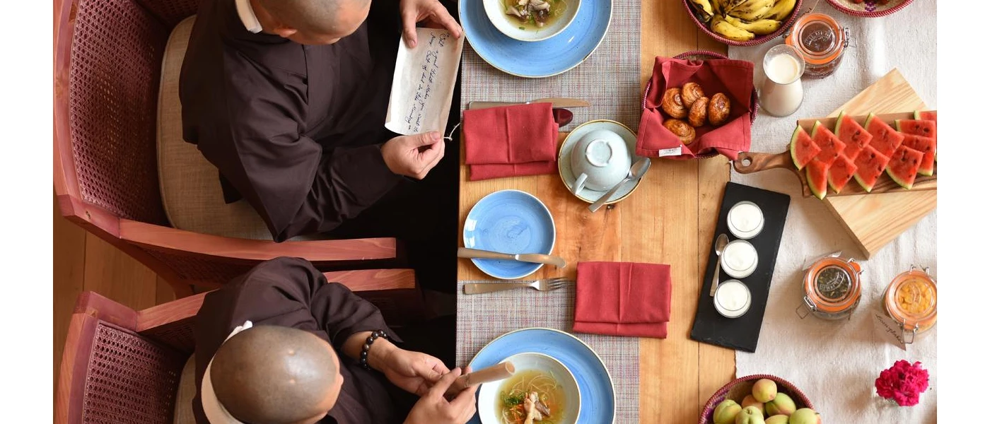 Two monks reading scripture while seated at a table laid out with pastries, watermelon and bananas
