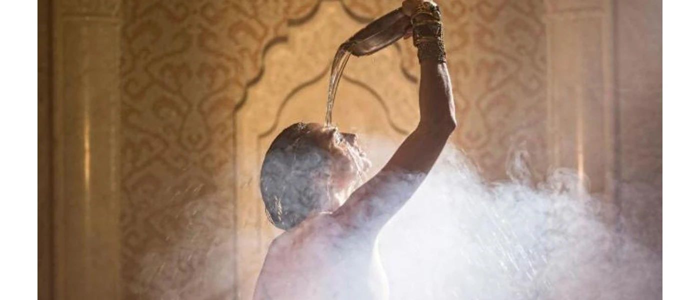 Woman pouring water over her head as steam rises around her in front of a Moroccan-style wall