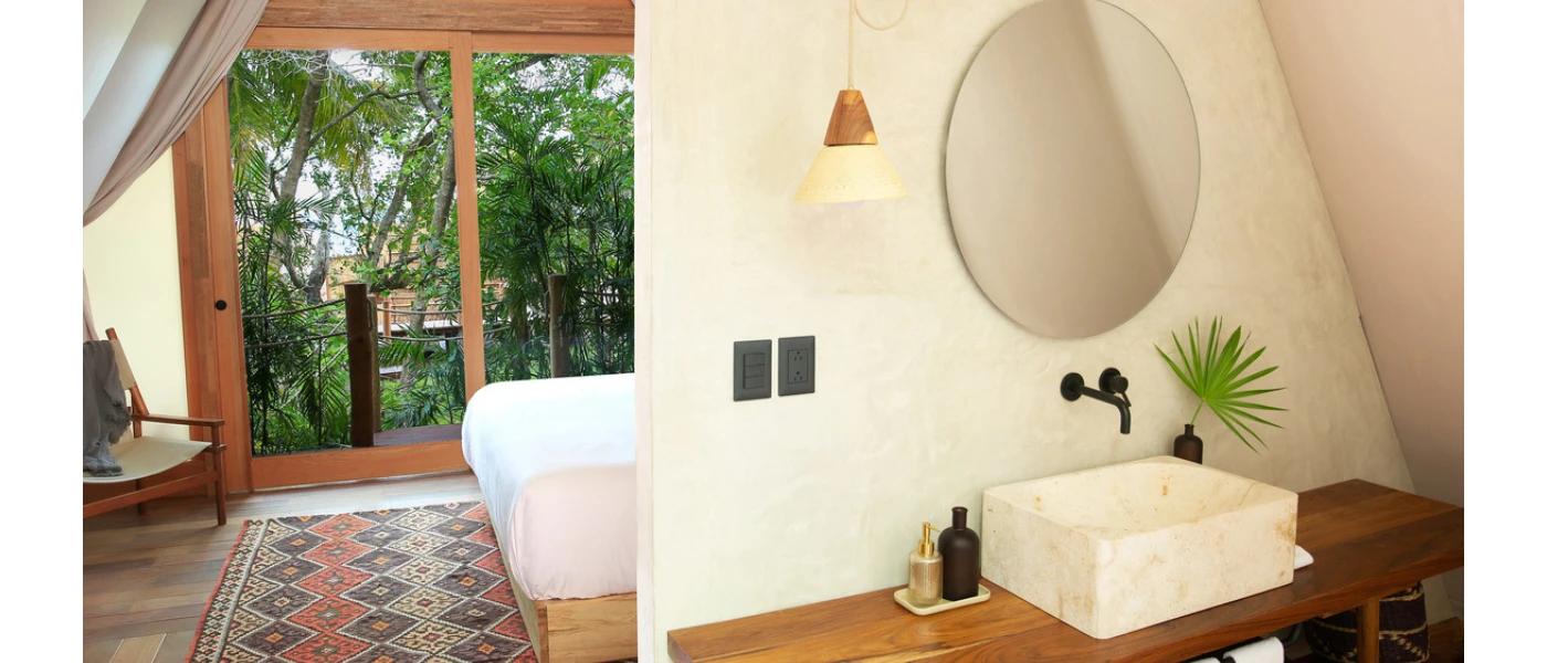 Airy bedroom and bathroom with a patterned rug, full-length balcony doors with a view of greenery, a wooden vanity unit and a white butler sink, plus a round mirror on the wall