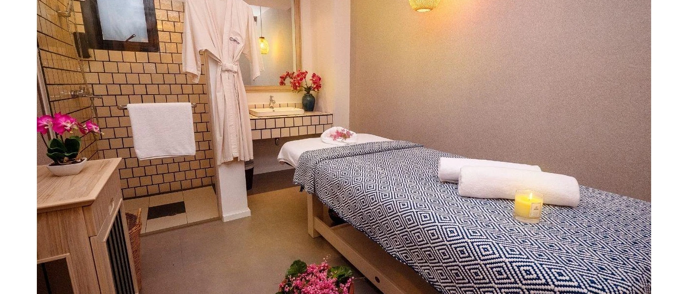 Treatment room with a cream tiled shower and vanity unit, a wooden side table, and massage bed with a blue blanket