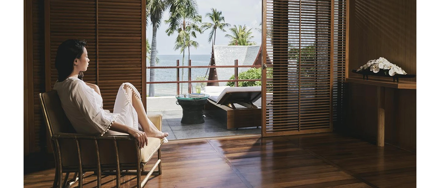 White-robed women sits in a hotel room decked out in dark woods, overlooking a furnished balcony and the palm trees and ocean in the distance