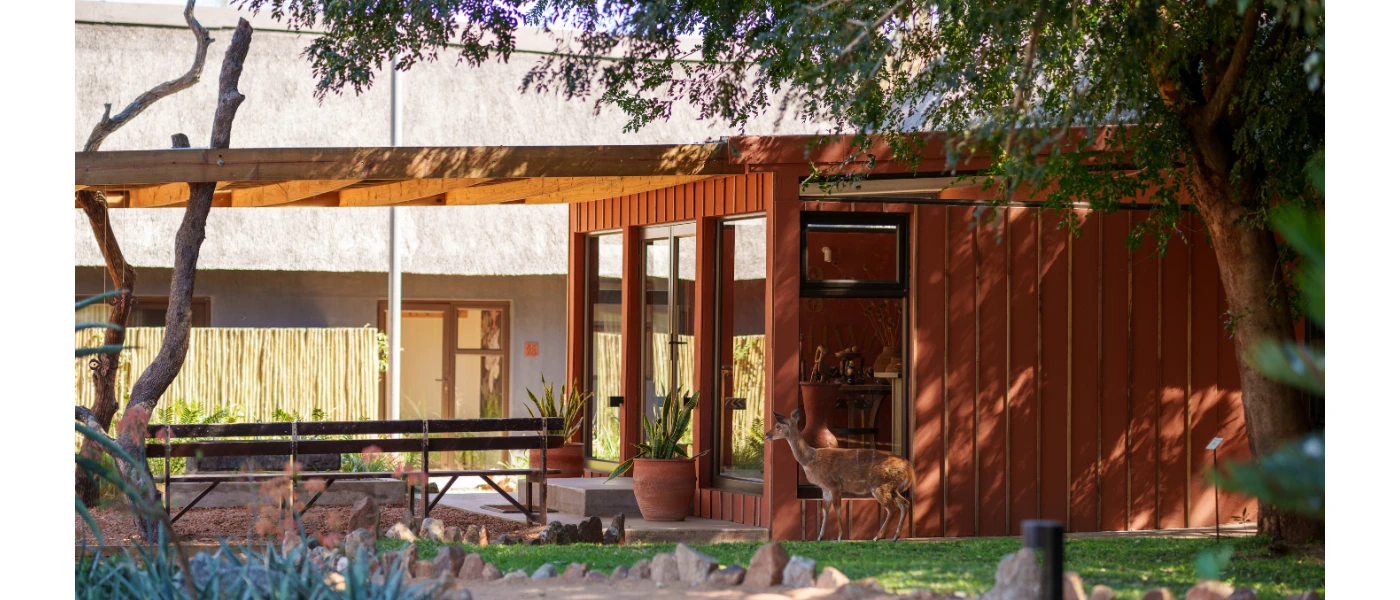 Low-rise terracotta building in a lawned area with trees, and a deer walking past