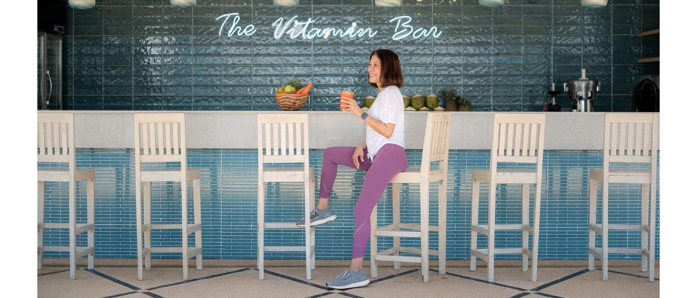 Smiling woman in purple leggings and a white T shirt sits at a bar sipping an orange smoothie or juice, with a sign behind her that reads 'The Vitamin Bar'