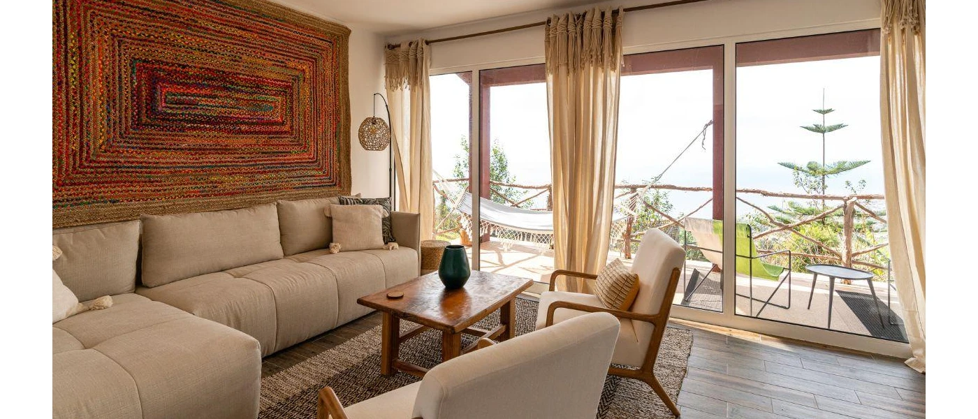 Beige cushions sofa on a jute rug next to a wooden coffee table, bohemian wall tapestry and light overlooking a countryside-facing terrace