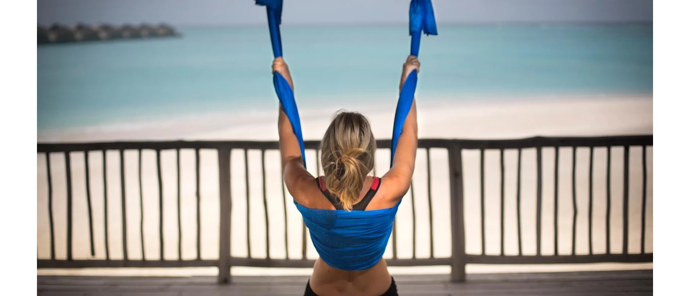 Woman in active wear hangs from blue ribbon on a terrace overlooking a white beach and turquoise shallows