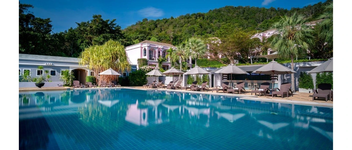 Swimming pool surrounded by white loungers and umbrellas, pink buildings and green hills