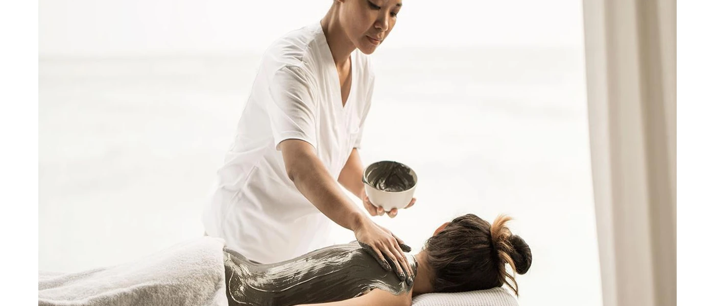Therapist applies mud to a woman's back as she lies on her front on a massage mus
