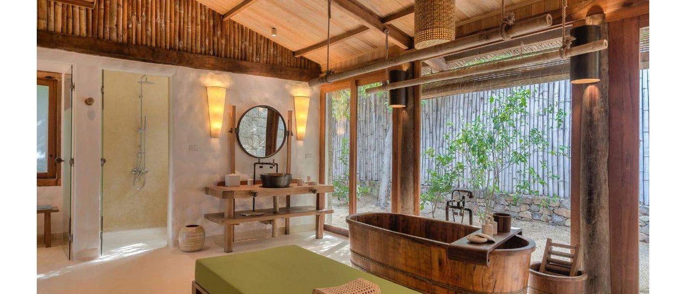 Villa bathroom with a wooden ceiling, vanity unit and bathtub, bohemian lighting and full-length windows overlooking leafy shrubs