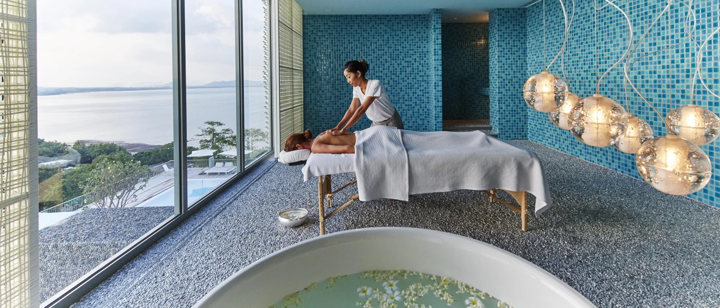 Therapist massages a woman on a bed in a room of blue mosaic tiles, with a round bath filled with white flowers and a large window with sea views
