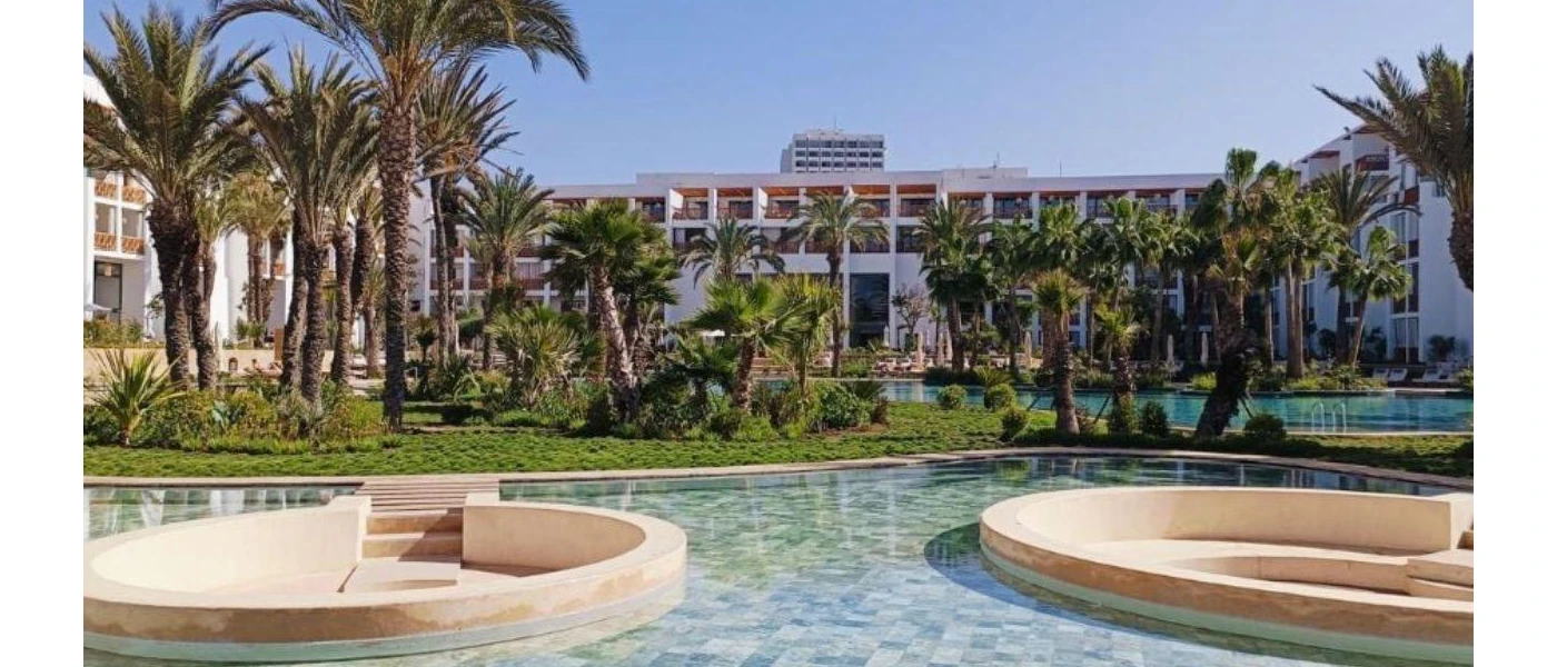 Outdoor pool and tropical greenery outside a high-rise white hotel under a blue sky