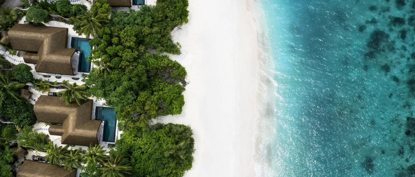 Aerial view of white sands, turquoise waters and tropical greenery, with thatched rooftops and swimming pools in between