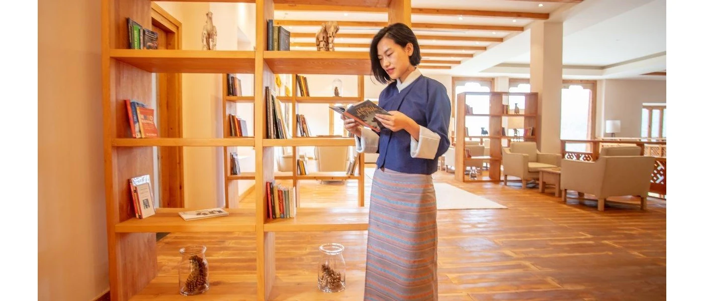 Woman in traditional dress reading a book next to a library shelf in a room with a wooden floor and wooden beams 