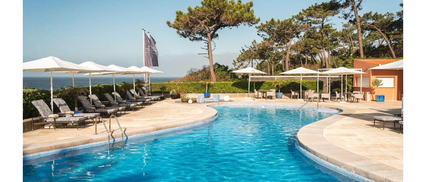 Curved swimming pool surrounded by white parasols, loungers with blue and white cushions and an ocean view