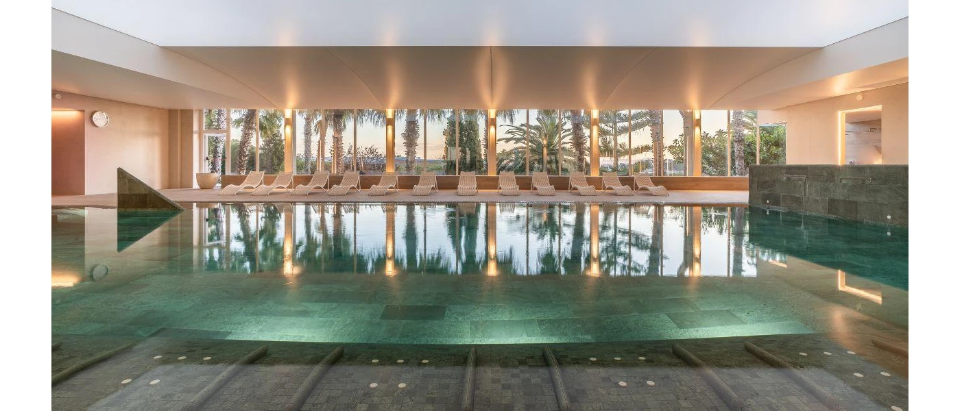Indoor pool with tiled flooring, surrounding loungers and full-length windows showcasing tropical grounds beyond