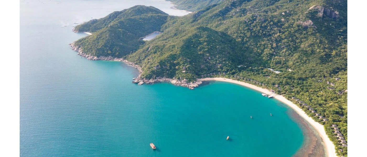 Aerial view of green hillsides, with a slice of white sand at the bottom and turquoise sea lapping the beach