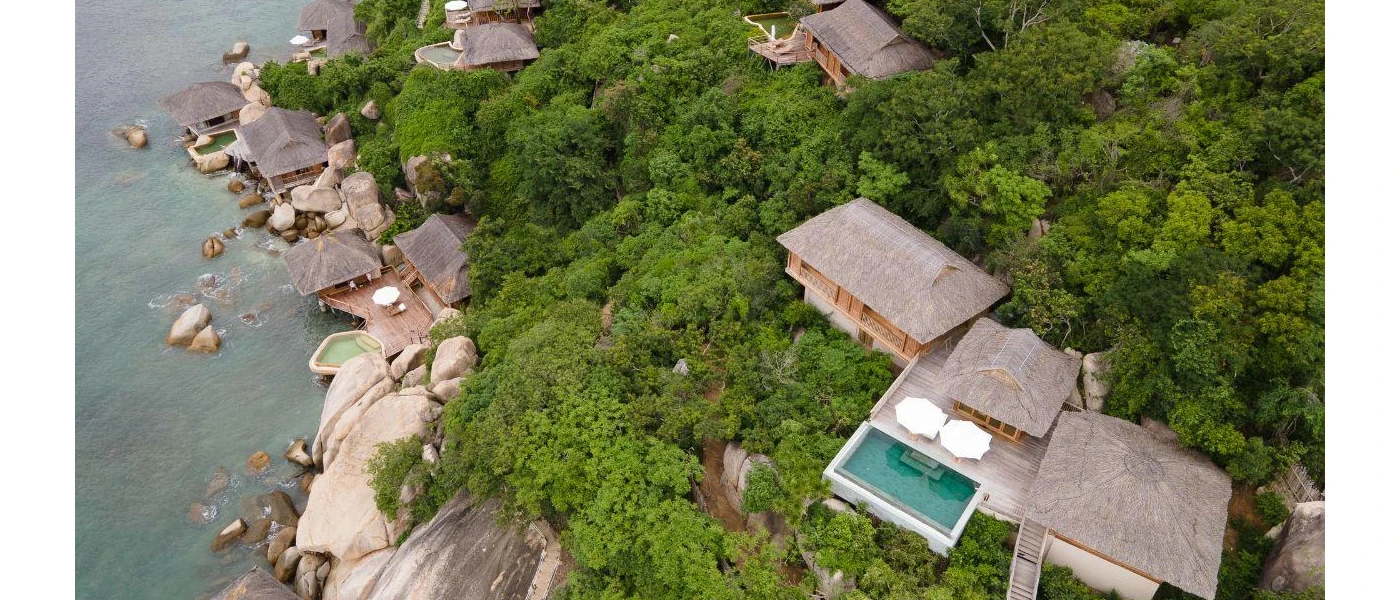 Thatched-roof villas with wooden decks and private pools nestled among thick greenery, next to striking rock formations and the sea, with the lowest villas showing off private pools with steps into the sea