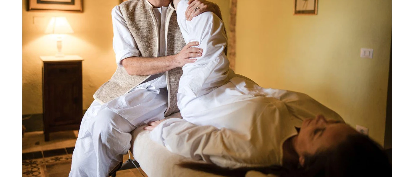 Woman in white lies on a bed as a therapist stretches one leg up into the air