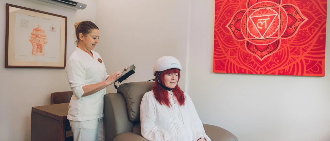 Woman sits with her eyes closed in a white helmet as a staff member reads from a digital device
