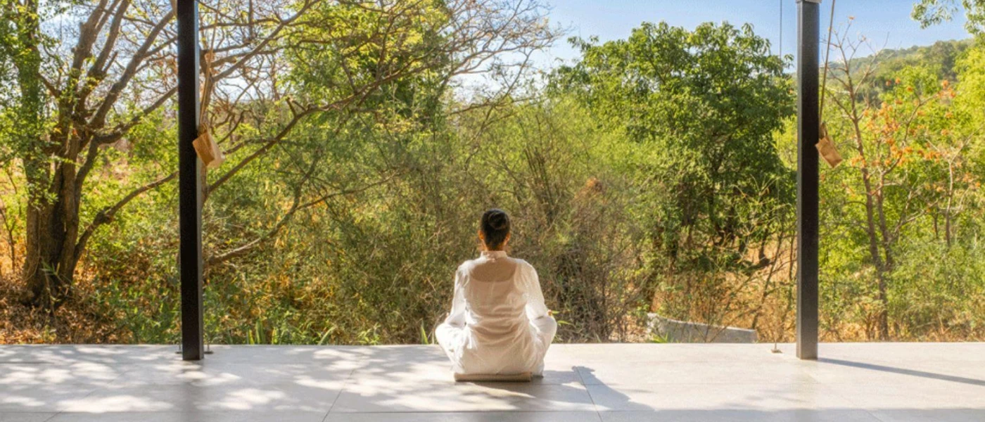 Woman sits cross-legged with her back to us in white clothes, overlooking surrounding forest from an open-sided pavilion